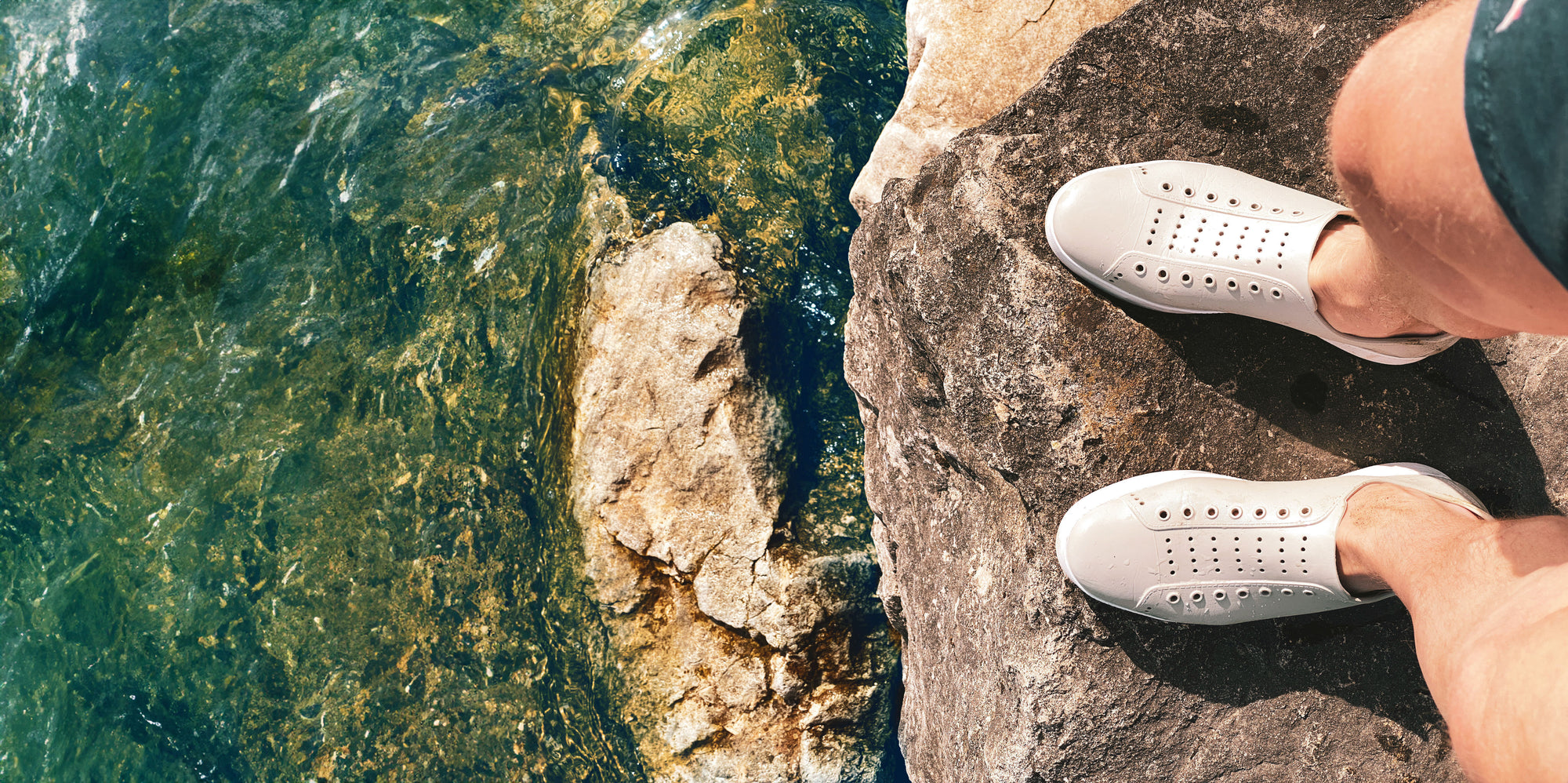 Person wearing white trainers, standing on a rock near a body of water