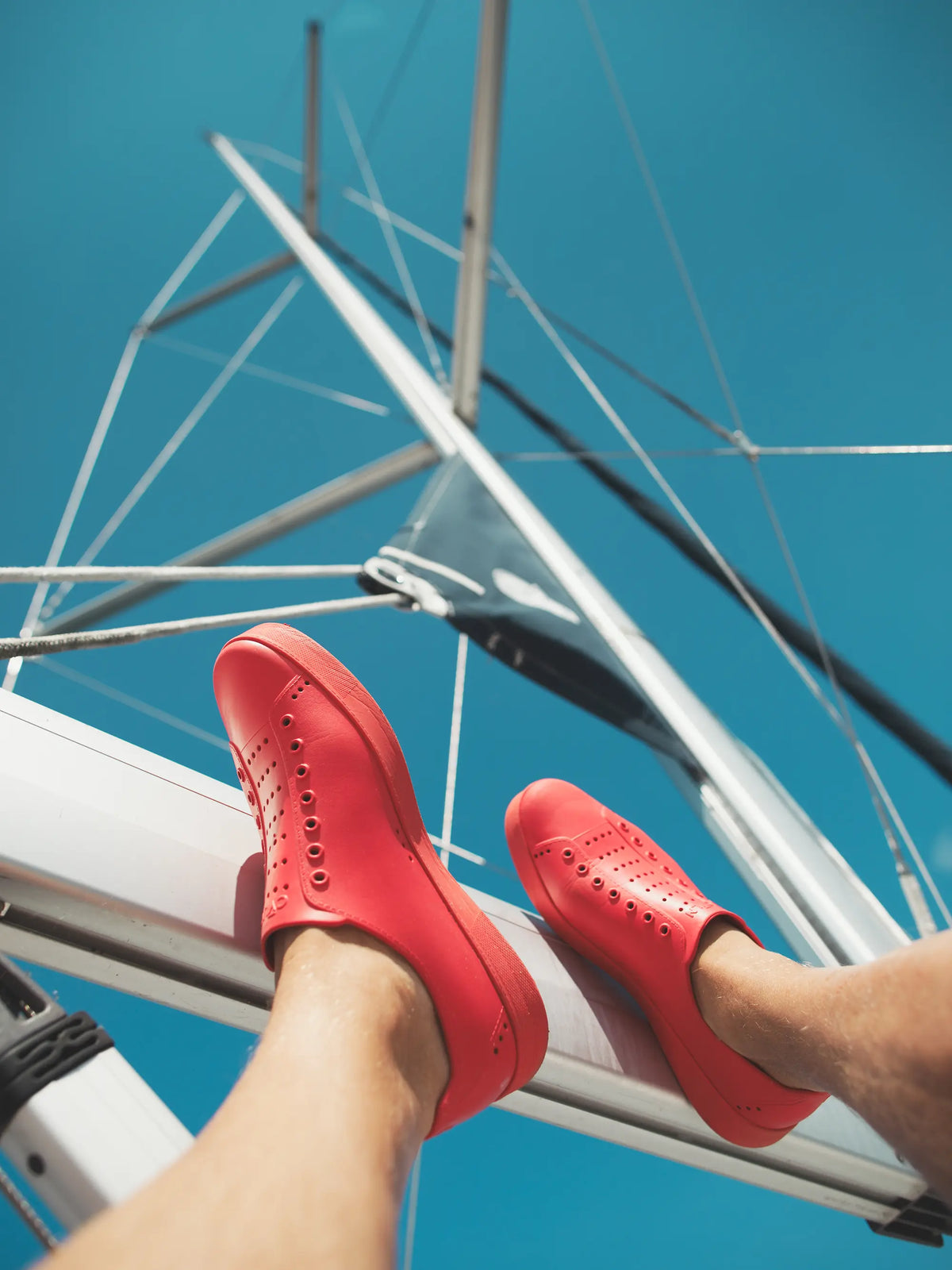 A person wearing bright red slip-on trainers, sitting on a white boat railing against a clear blue sky and sailboat mast.